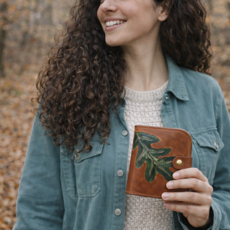 Mujer sosteniendo una billetera artesanal de cuero marrón con una hoja de roble grabada y pintada a mano sobre piel natural.