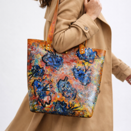 Mujer llevando bolso tote artesanal de cuero pintado a mano con flores azules sobre fondo multicolor.